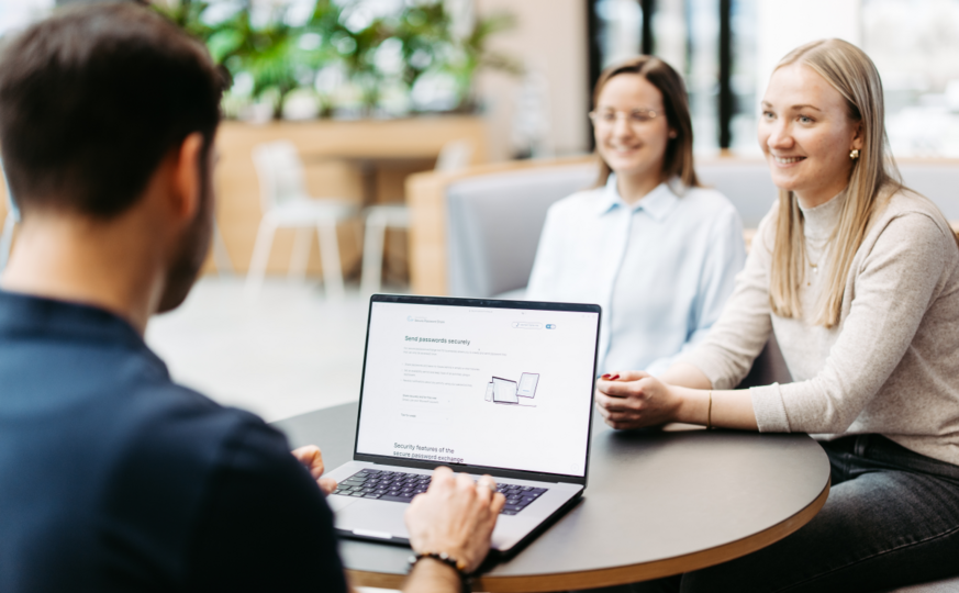 Drei Personen sitzen an einem Tisch und besprechen sich. Wir schauen einem Mann über die Schulter. Er hat einen Laptop vor sich und nutzt Secure Password Share.